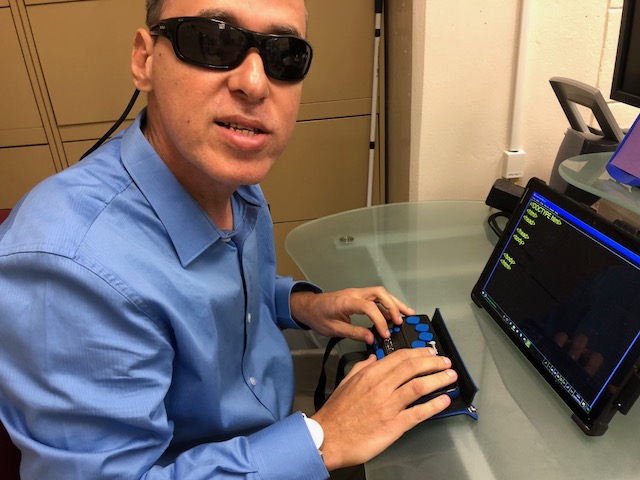 La foto presenta a Manolo en su escritorio programando con su línea braille frente a su computadora. Foto de Manolo sonriendo utilizando una computadora con una línea Braille en su escritorio.