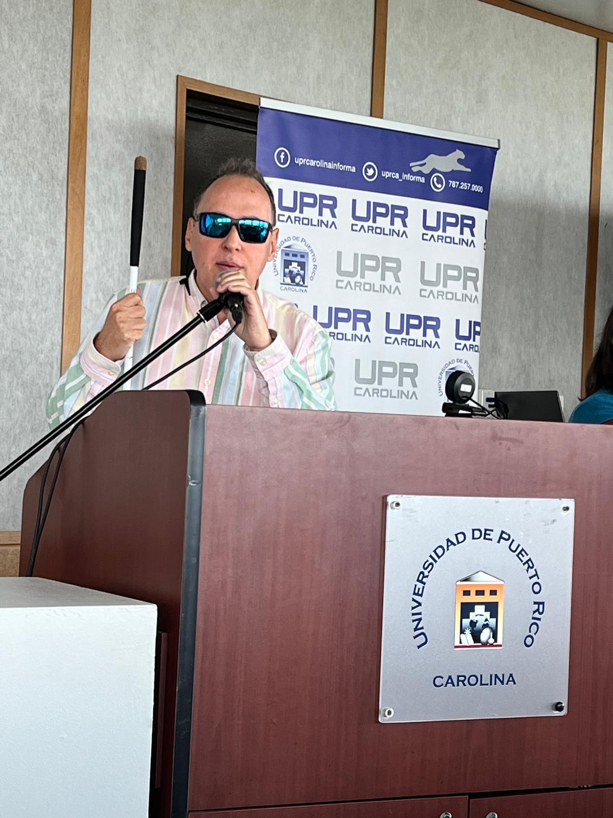 La foto presenta a Manolo y se observa el cartel de la Universidad de Puerto Rico, Recinto de Carolina. Foto de Manolo en el podio haciendo su presentación.