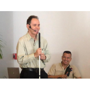 La foto presenta a Manolo de pie y sujetando su bastón blanco posando con el maestro de ceremonias de la actividad que se encuentra en silla de ruedas.