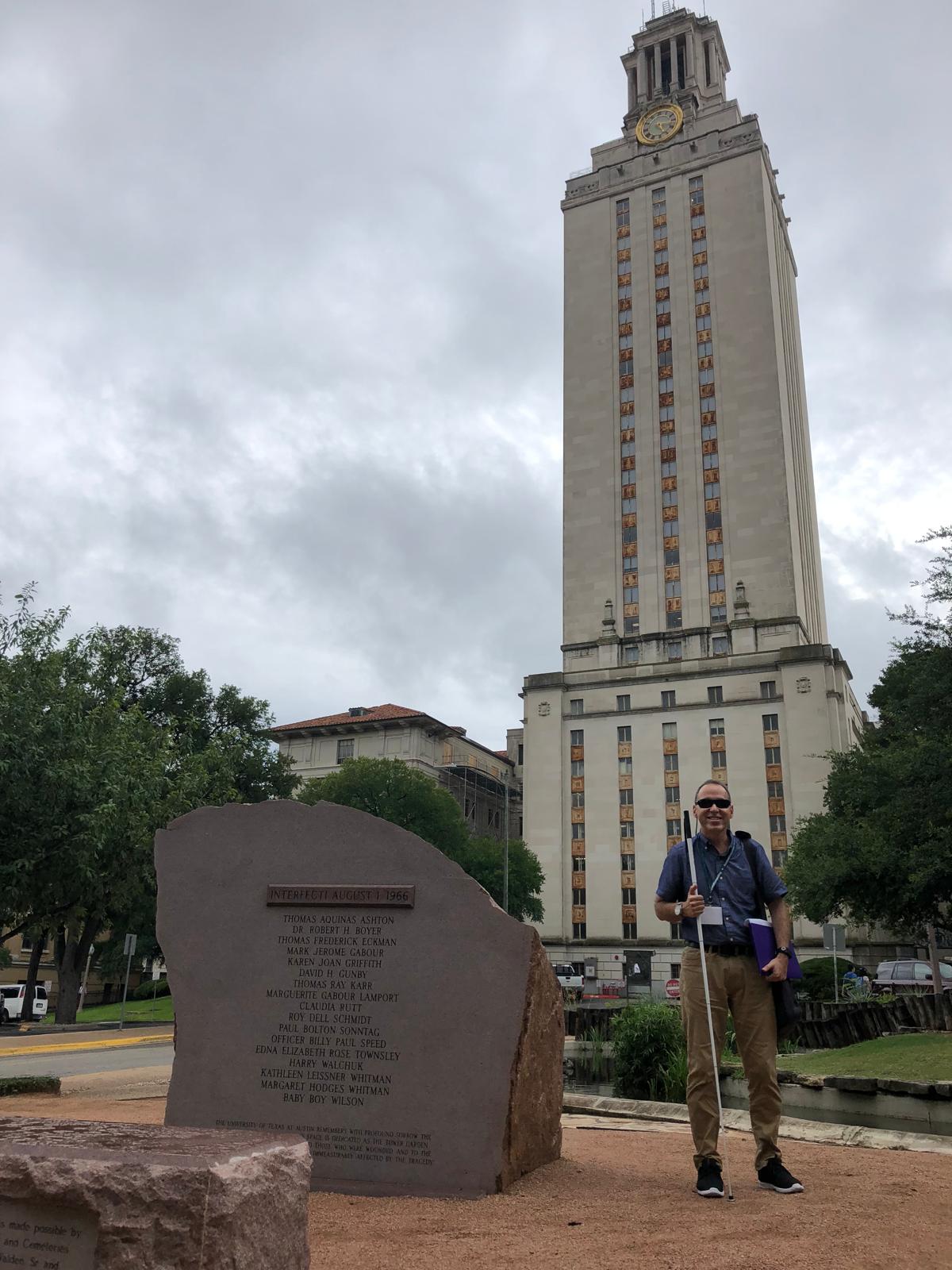 Manolo tiene en una mano sujetado su bastón blanco y en la otra mano una carpeta. Foto de Manolo posando al lado de una torre alta con un reloj en la parte superior, que es el icónico edificio de la Universidad de Texas en Austin.