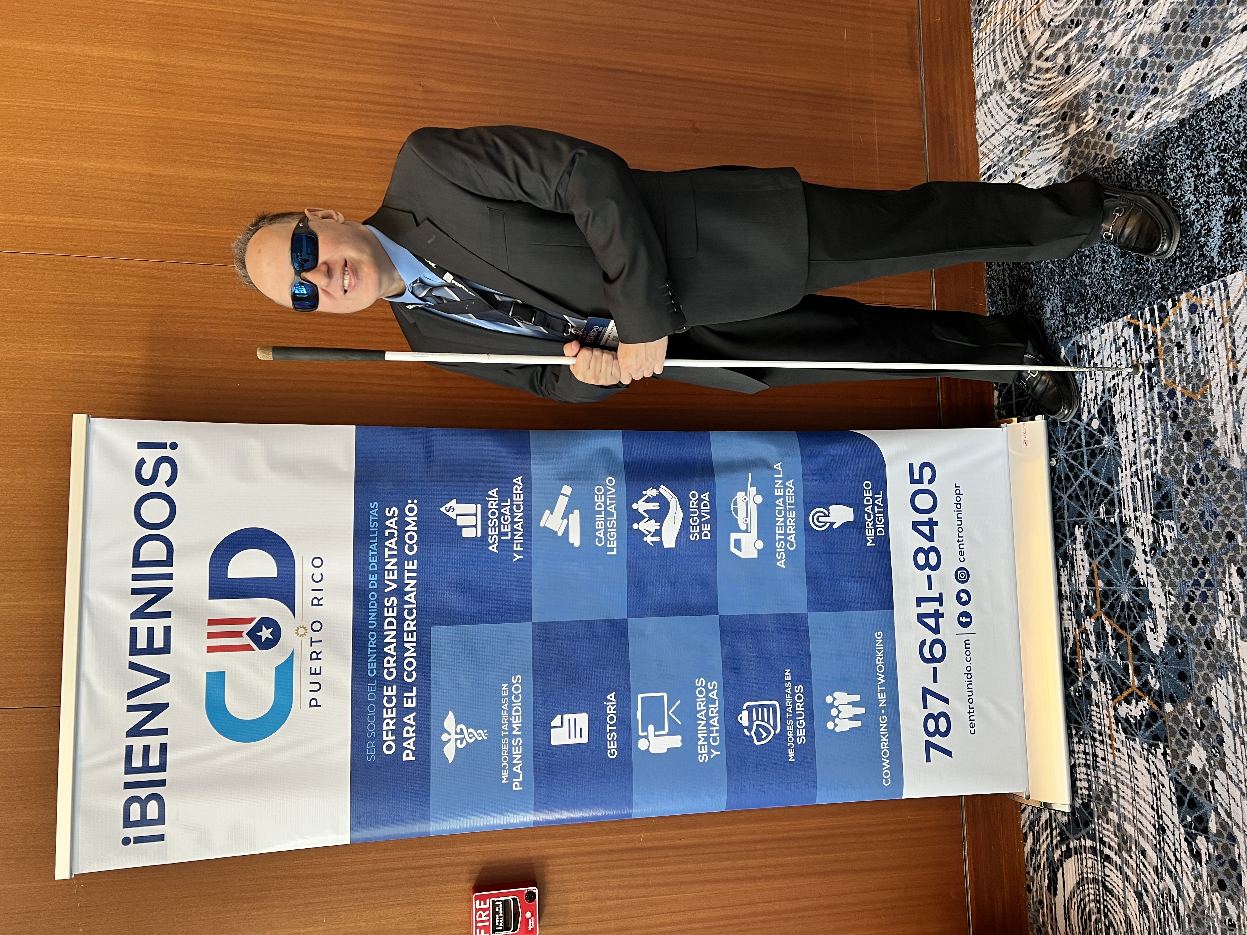 La foto representa la participación de Manolo en la conferencia del Centro Unido de Detallistas de Puerto Rico. La foto muestra a Manolo de pie junto a un cartel. Manolo lleva un traje oscuro, una camisa azul y una corbata, y está usando gafas de sol. Sostiene un bastón blanco en su mano derecha. El cartel a su lado tiene un fondo blanco y azul, y en la parte superior dice