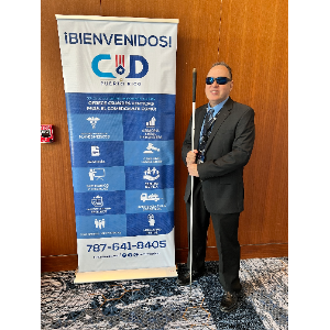 La foto muestra a Manolo de pie junto a un cartel. Manolo lleva un traje oscuro, una camisa azul y una corbata, y está usando gafas de sol. Sostiene un bastón blanco en su mano derecha. El cartel a su lado tiene un fondo blanco y azul, y en la parte superior dice 