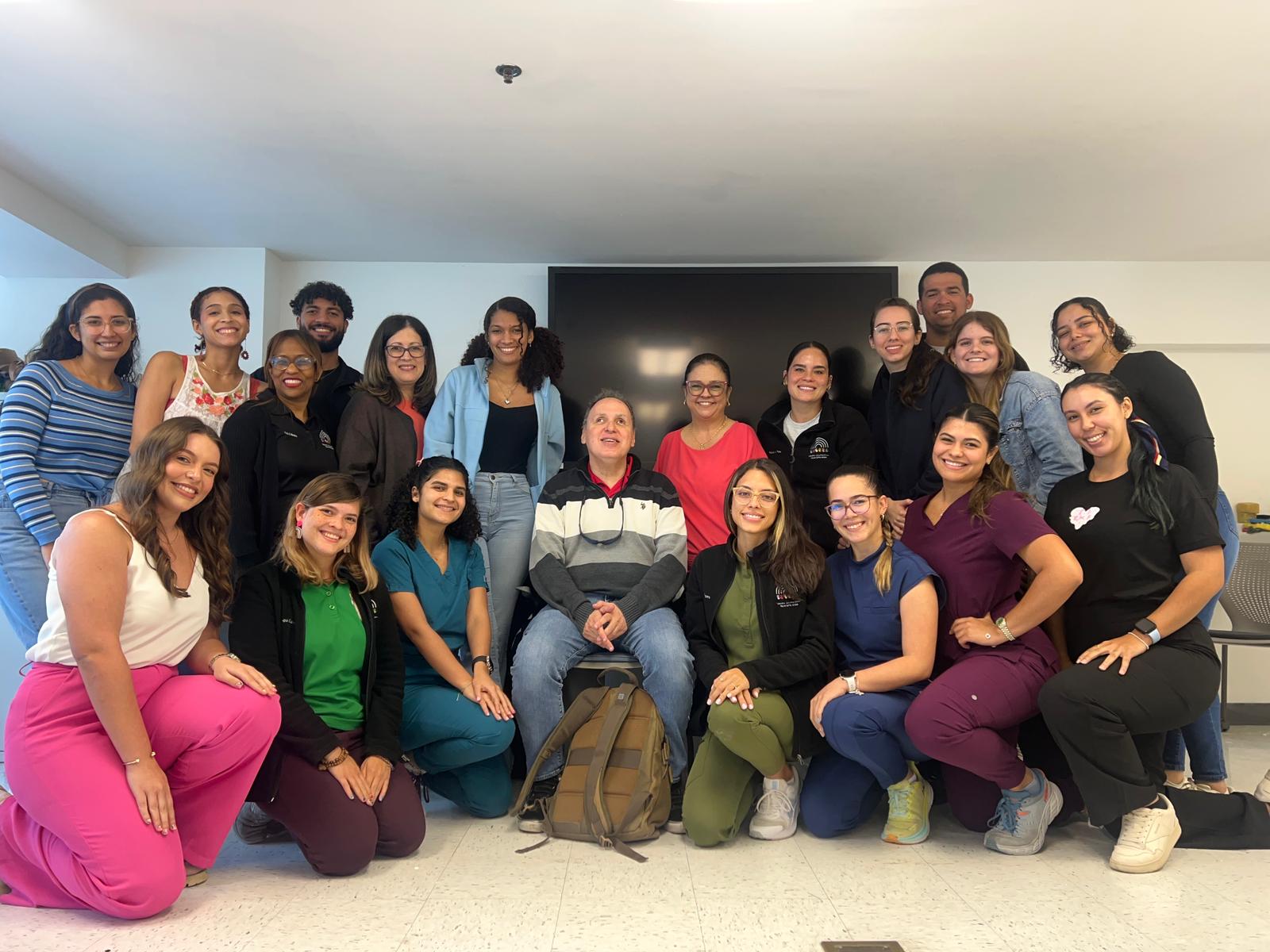 La foto representa la charla de Manolo a los estudiantes de maestría de terapia ocupacional del Recinto de Ciencias Médicas. La foto presenta a Manolo sentado en el centro de un grupo de estudiantes de terapia ocupacional. Hay estudiantes que están de pie y otros de rodillas posando para la foto.