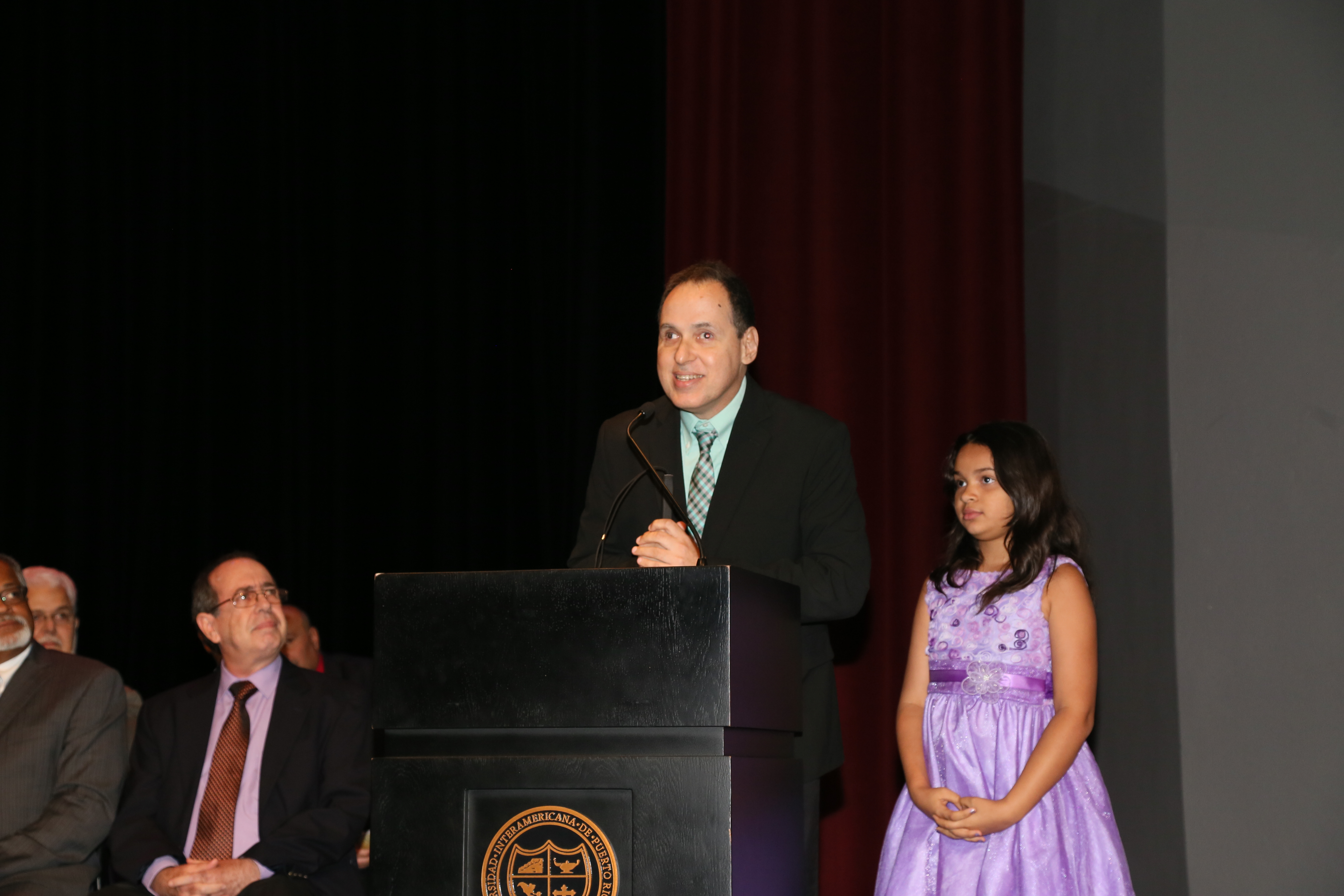 El podio dice en la parte de alfrente Escuela de Derecho de la Universidad Interamericana. La foto muestra a Manolo junto con su hija Ámbar mientras ofrecen un mensaje detrás del podio.