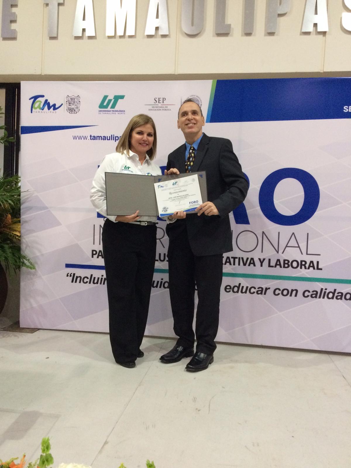 En el fondo se pueden ver los logotipos de la Universidad Tecnológica de Tamaulipas Norte. La foto muestra a Manolo recibiendo un certificado de una mujer en la tarima. La mujer a la izquierda lleva una camisa blanca con logotipos y sostiene un diploma o certificado. Mientas que Manolo está a la derecha y viste un traje oscuro con una corbata y también sostiene el diploma.