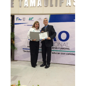 La foto muestra a Manolo recibiendo un certificado de una mujer en la tarima. La mujer a la izquierda lleva una camisa blanca con logotipos y sostiene un diploma o certificado. Mientas que Manolo está a la derecha y viste un traje oscuro con una corbata y también sostiene el diploma.