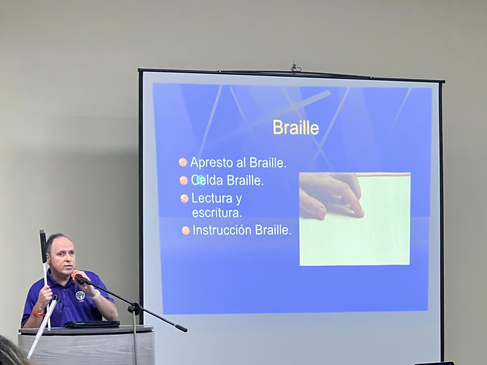 La foto muestra a Manolo en la parte frontal y una pantalla con la presentación en Power Point con el tema del Braille y la tecnología. La foto presenta a Manolo en el podio durante su charla.