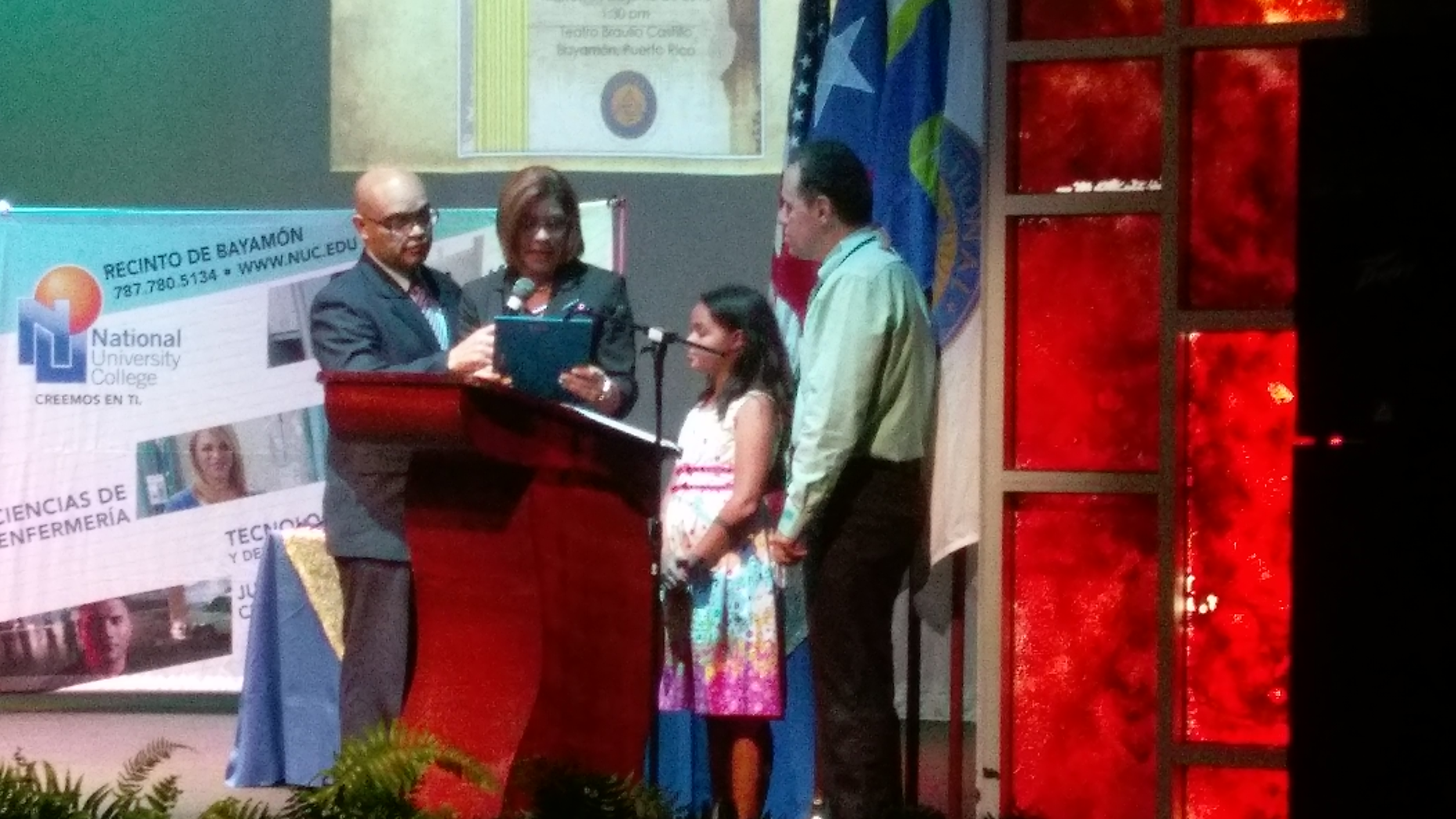 En la parte de atrás de la tarima, se observa una pantalla con información para los graduandos. La foto muestra a Manolo junto con su hija Ámbar como parte del mensaje en la ceremonia al lado del podio.