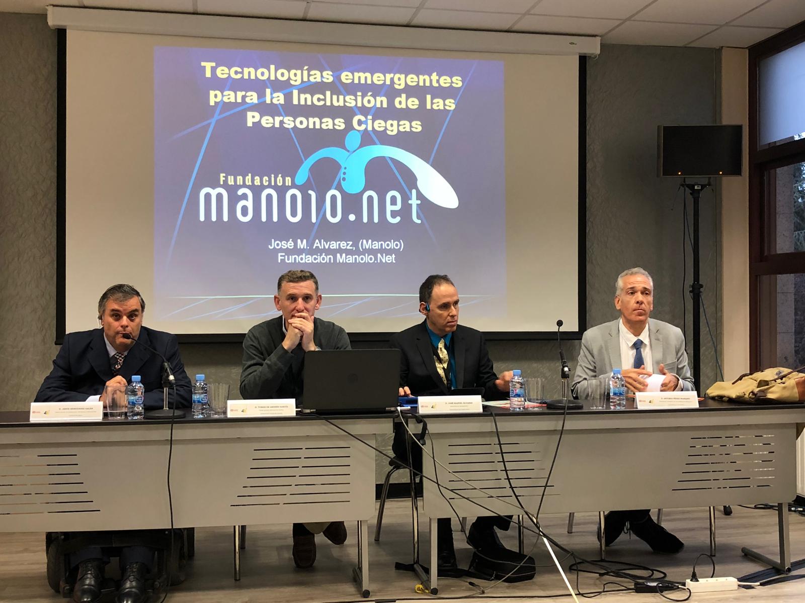 Los cuatro hombres están vestidos formalmente y hay micrófonos y botellas de agua sobre la mesa. La foto muestra a cuatro hombres sentados detrás de una mesa durante la conferencia CIUD 2018 en España. Detrás de ellos hay una pantalla de proyección que muestra el título Tecnologías emergentes para la Inclusión de las Personas Ciegas.