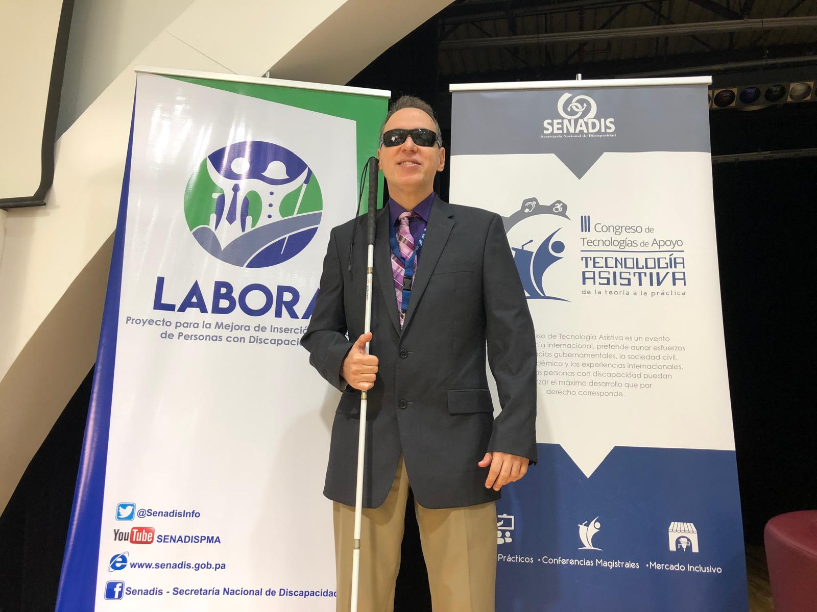 La pancarta a la izquierda tiene el logo de LABORA y el texto La foto muestra a Manolo vestido con un traje oscuro, camisa morada y corbata de colores, sosteniendo su bastón blanco. A su lado hay dos pancartas que anuncian la conferencia en Panamá.