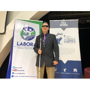 La foto muestra a Manolo vestido con un traje oscuro, camisa morada y corbata de colores, sosteniendo su bastón blanco. A su lado hay dos pancartas que anuncian la conferencia en Panamá.
