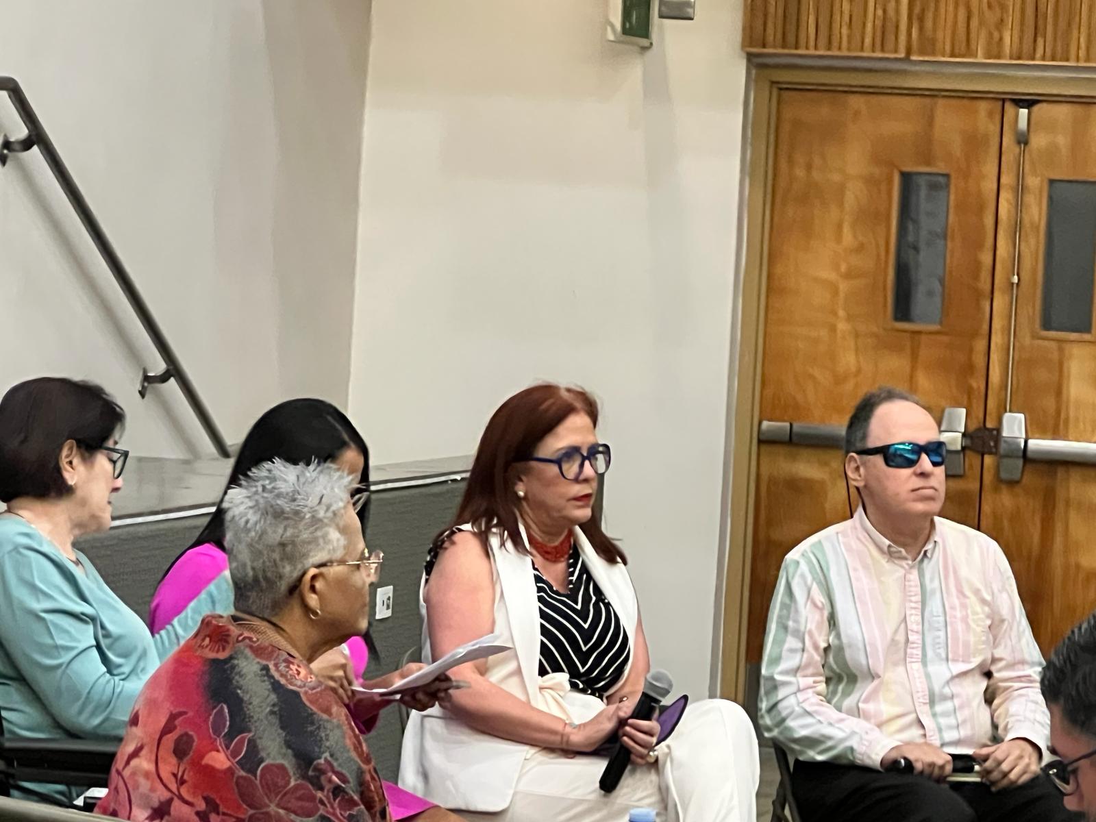 La foto presenta a los cinco panelistas sentados en la tarima. Manolo tiene una camisa de manga larga color pastel y tiene puesta gafas de sol. Foto de Manolo como parte del panel.