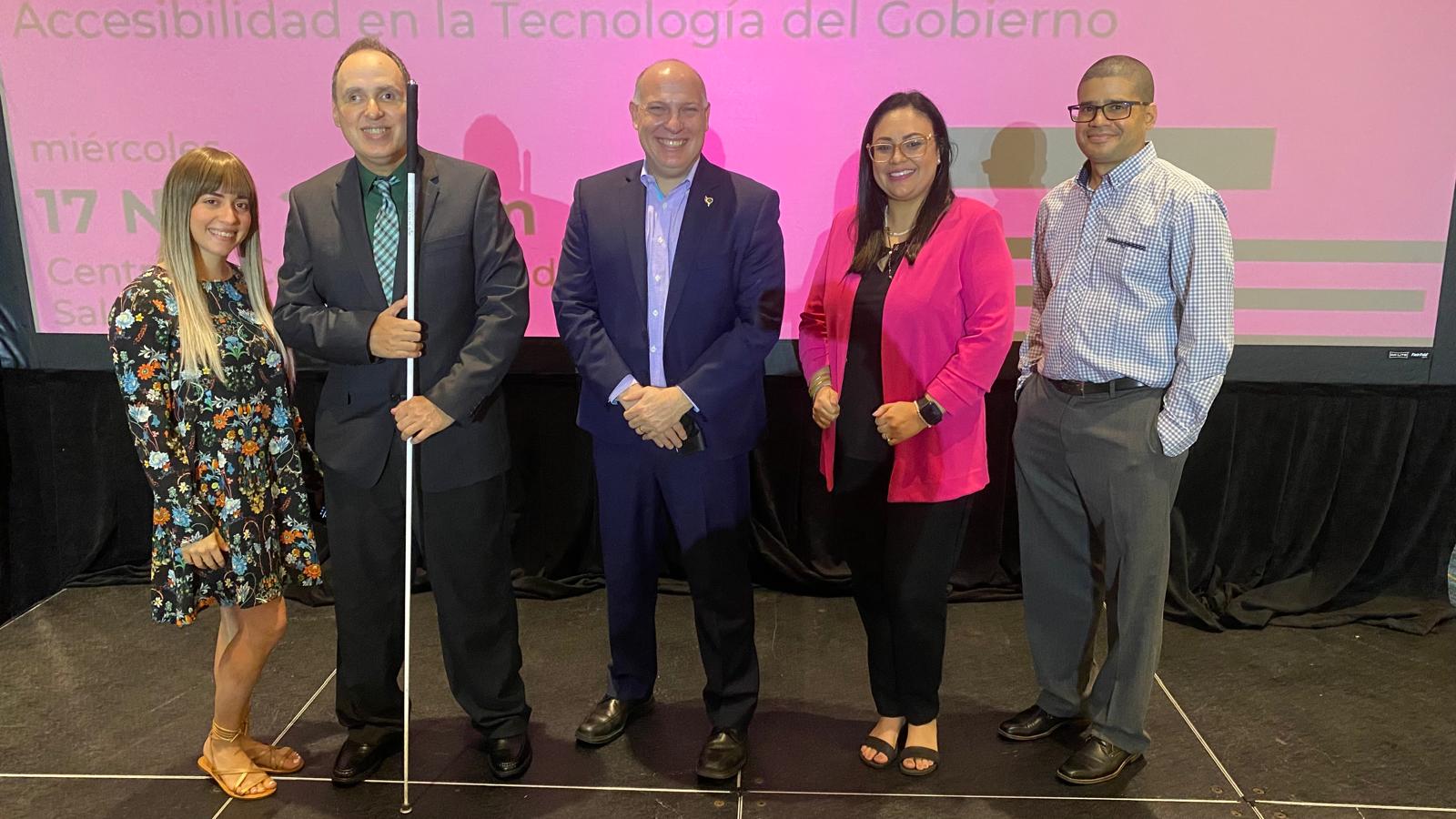 La foto presenta a Manolo en la tarima detrás del podio, se observa la pantalla con su presentación y al público presente en la actividad. Foto de Manolo durante su presentación en el evento.