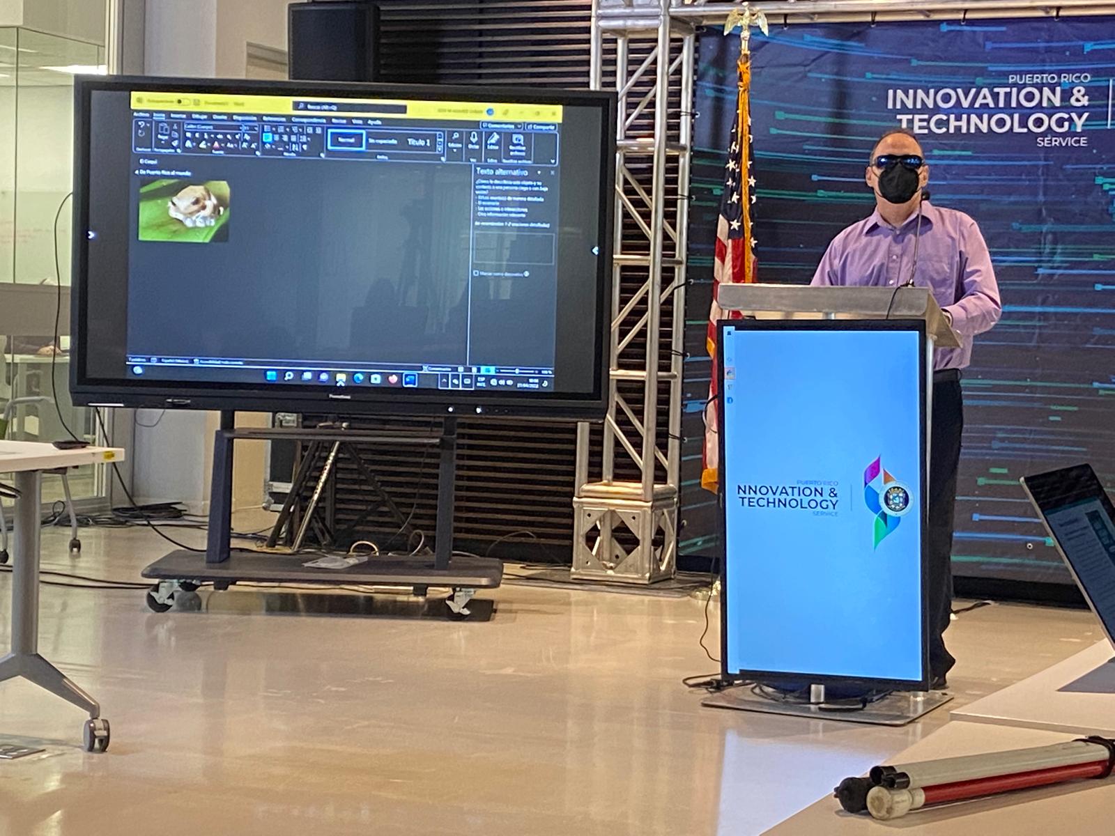 La foto muestra la pantalla con la presentación en la parte de atrás. Foto de Manolo haciendo su presentación utilizando su computadora en el podio.