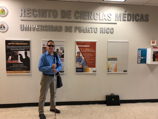 Manolo se encuentra de pie y tiene sujetado su bastón blanco. La foto presenta a Manolo posando al lado del letrero del Recinto de Ciencias Médicas de la UPR.