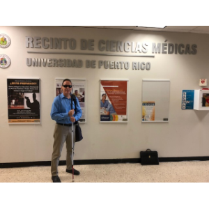 La foto presenta a Manolo posando al lado del letrero del Recinto de Ciencias Médicas de la UPR.