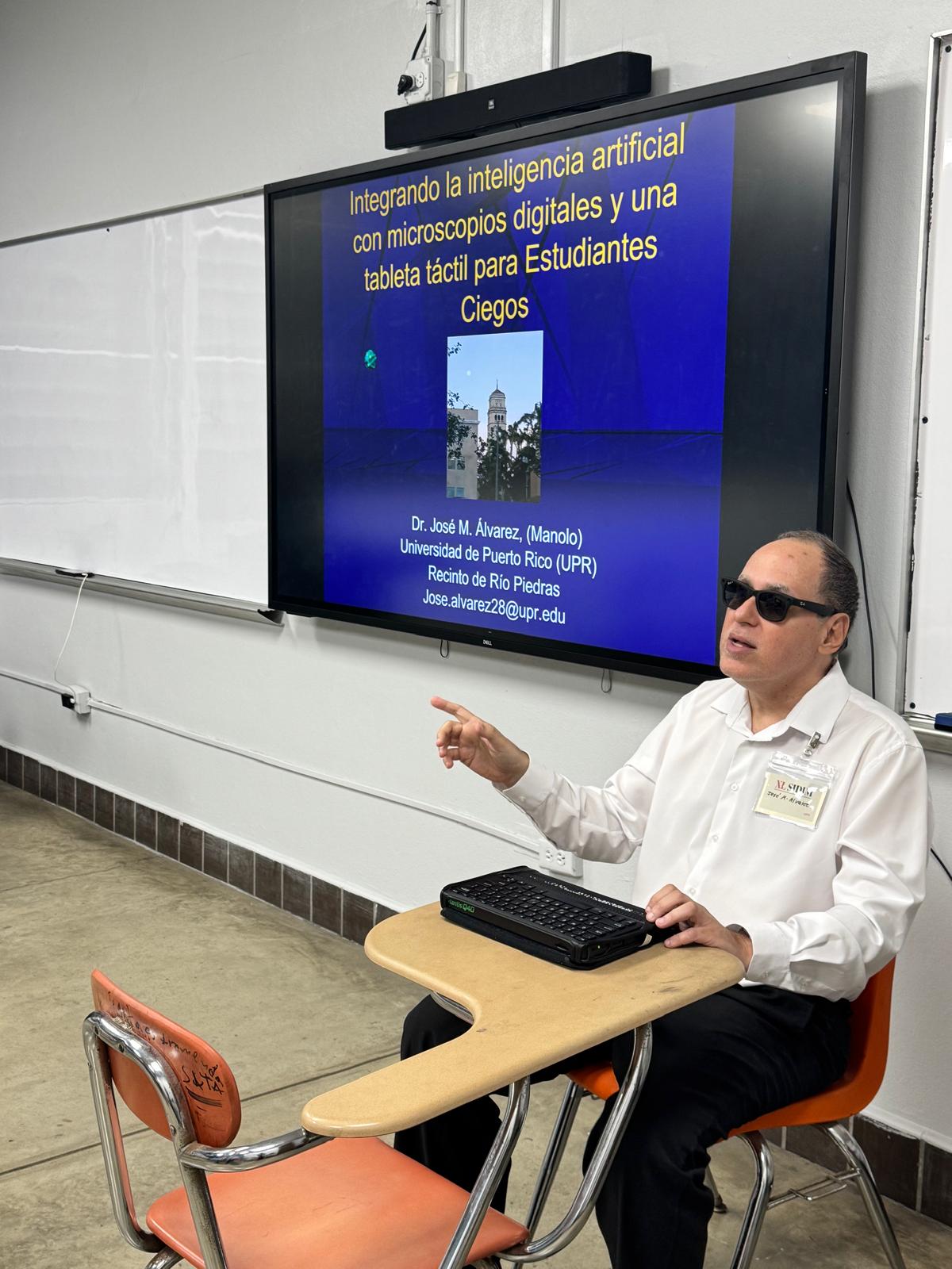 La foto muestra también al lado de Manolo una pantalla donde se observa parte de su presentación en Power Point. La foto muestra a Manolo sentado en la parte frontal del salón durante su presentación.