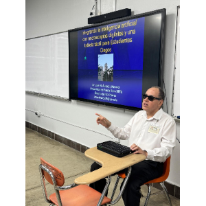 La foto muestra a Manolo sentado en la parte frontal del salón durante su presentación.