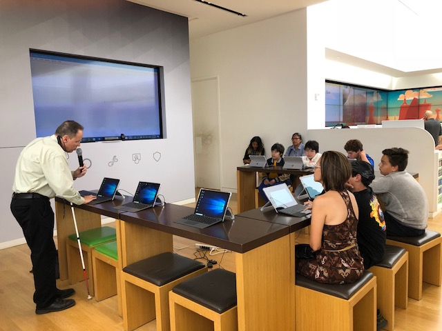 La foto presenta el área central del teatro de la tienda de Microsoft. Foto de Manolo junto a varios participantes del taller.