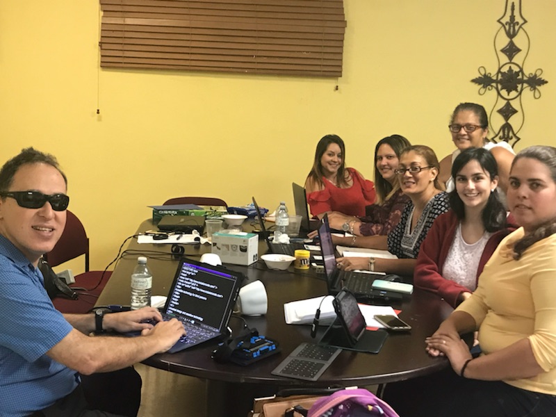 La foto muestra a Manolo utilizando su computadora y a un grupo de maestras de educación especial, programando en sus respectivas computadoras. Todos están en una misma mesa. Foto de Manolo enseñando programación a grupo de maestras de educación especial.